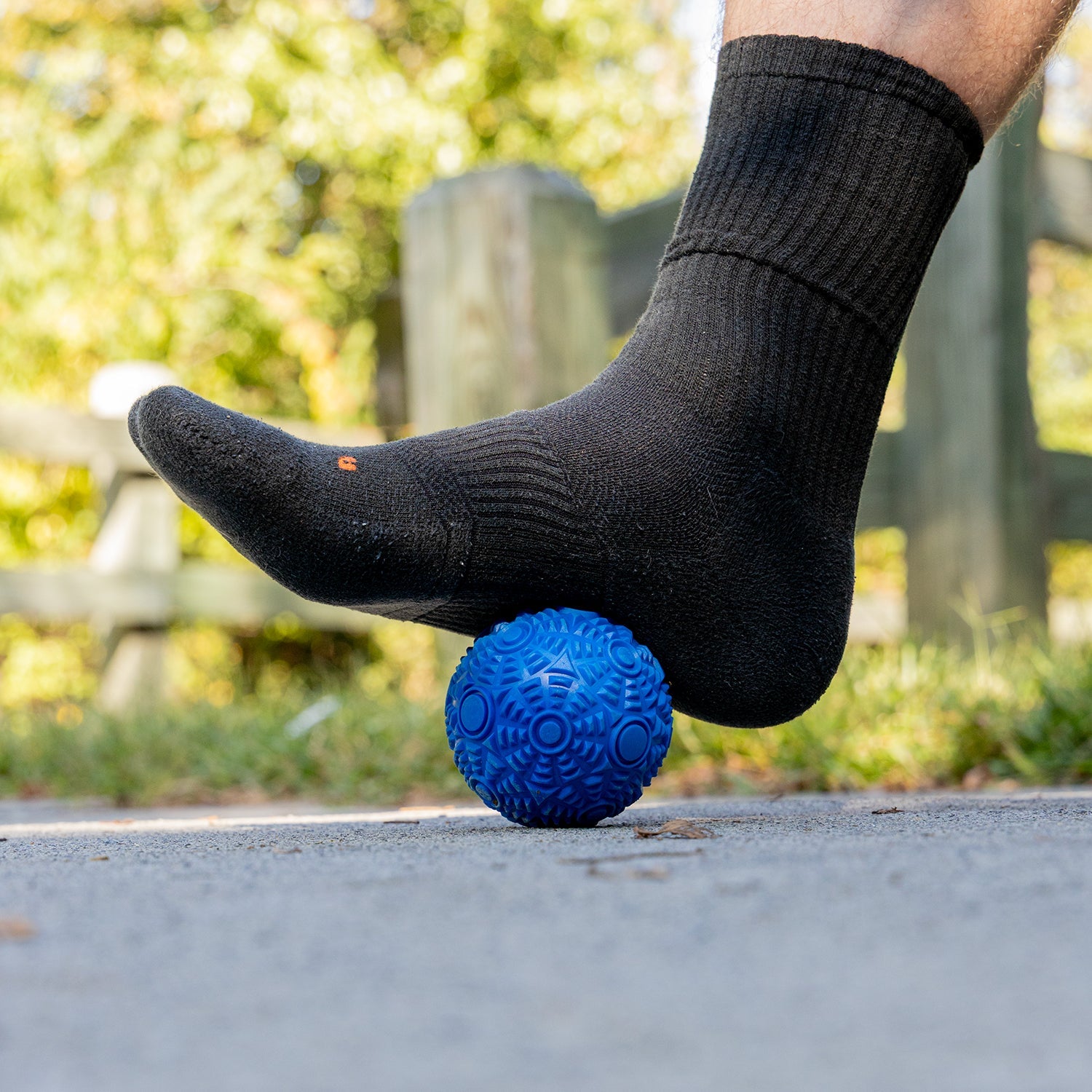 A person wearing a white sock with a black Nike logo rolls the bottom of their foot on a red massage ball on a black textured floor.