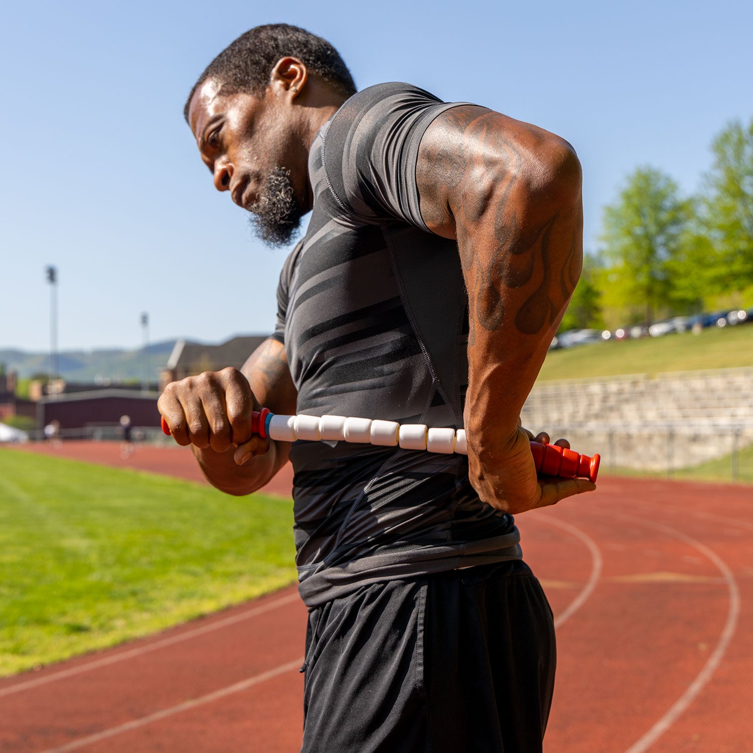 A man in athletic wear uses the Travel Stick-18 on his back for muscle recovery while standing on an outdoor running track on a sunny day.