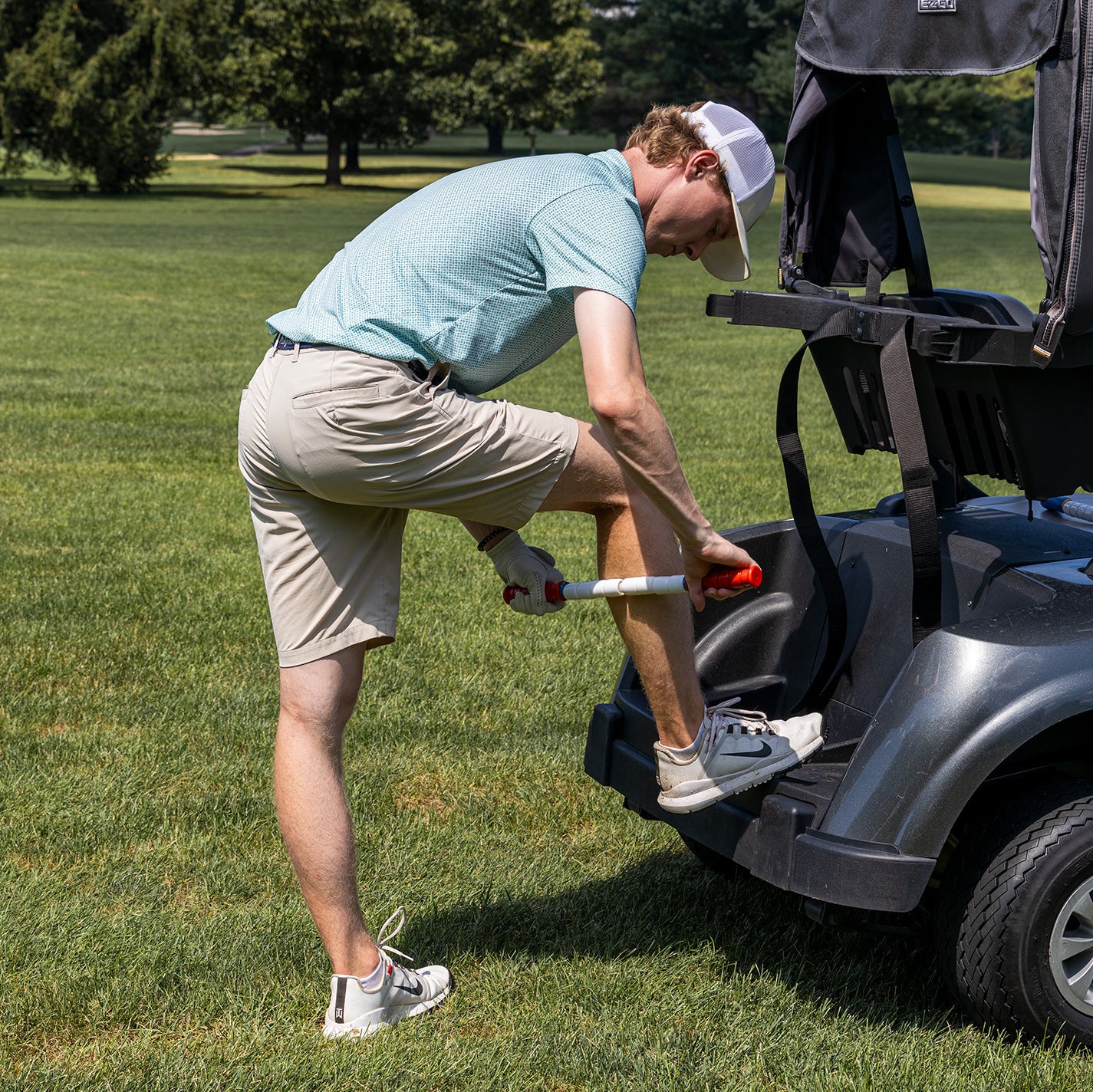 A man in a light blue polo, khaki shorts, and white cap stands by a golf cart on a grassy course, using his Travel Stick-18—a gym bag essential—to massage his right calf for muscle recovery.