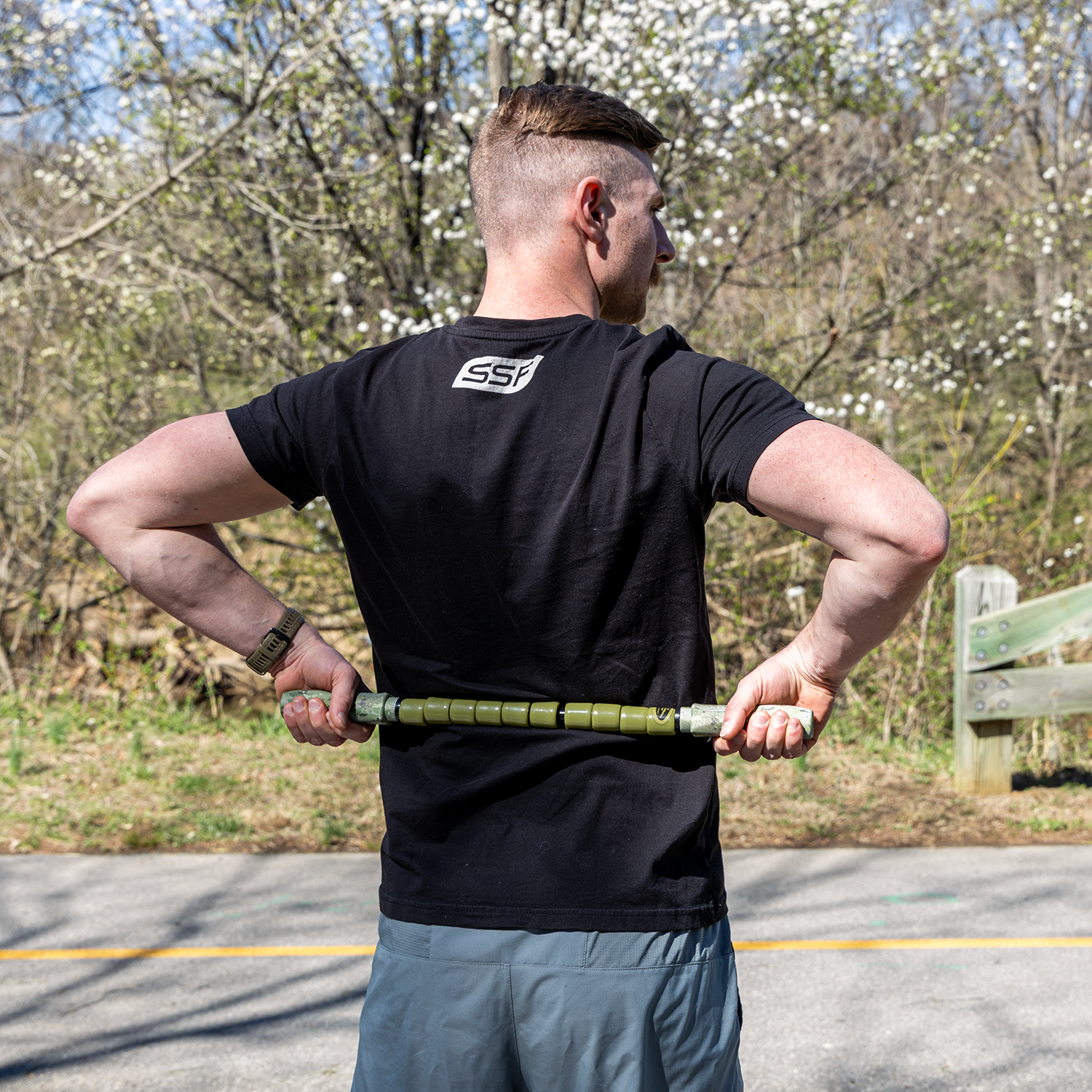A person in a black T-shirt and gray shorts uses the Hunter Stick- 22 on their lower back while standing near a road, surrounded by trees and blooming flowers—ideal for muscle relief during adventure travel.
