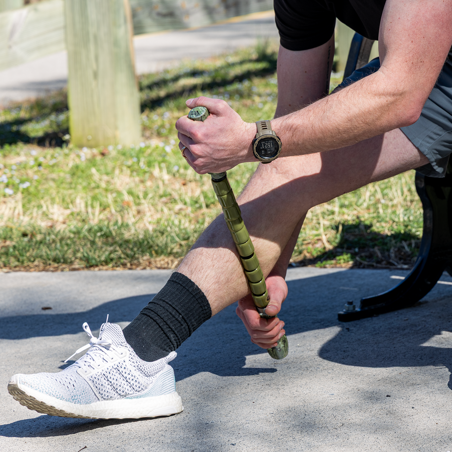 A person sits on a bench outdoors using the Hunter Stick-22 to roll their lower leg for muscle relief. They wear a black shirt, gray shorts, black socks, white sneakers, and a smartwatch after an adventure.