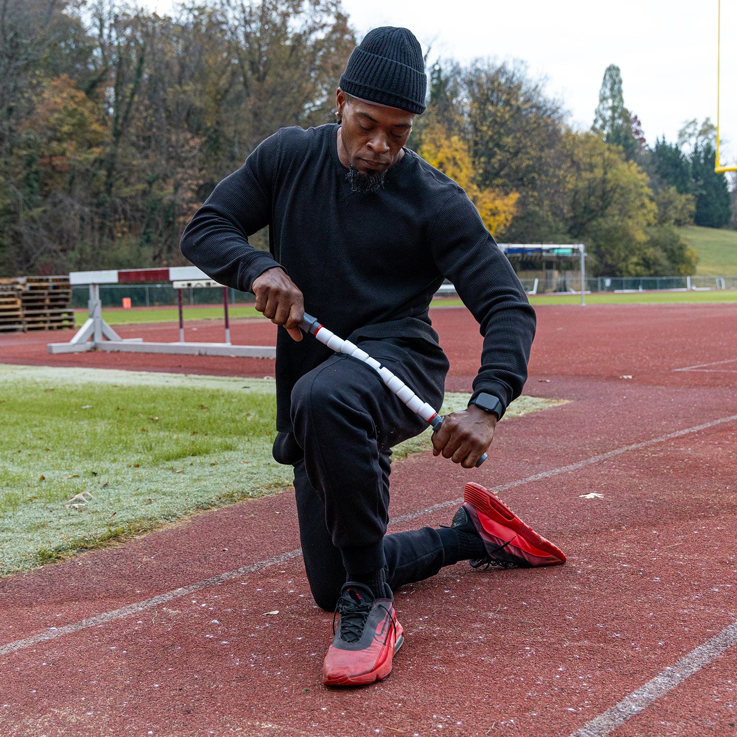 Man wearing all black kneeling on an outdoor track using the Sprinter Stick to massage his outer quad.