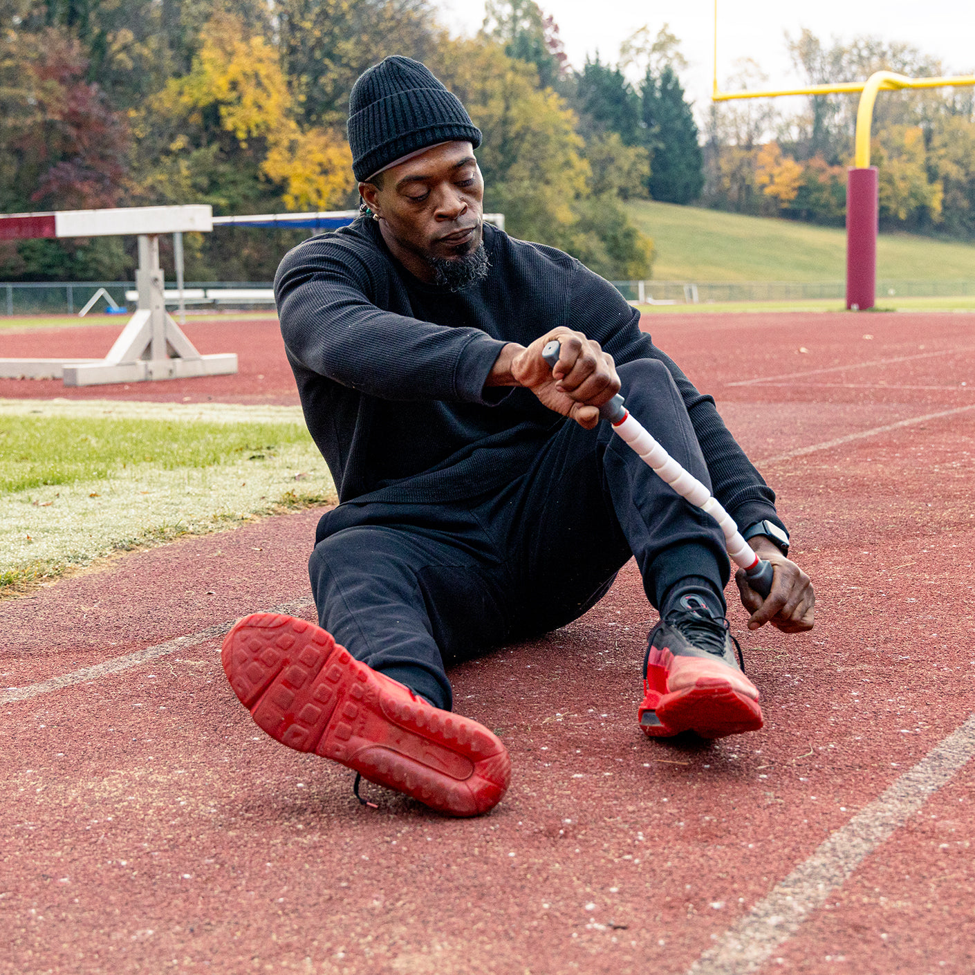 A man in black athletic wear sits on an outdoor track, stretching his leg using the Sprinter Stick- 22 for muscle recovery. Autumn trees and sports equipment are visible in the background.