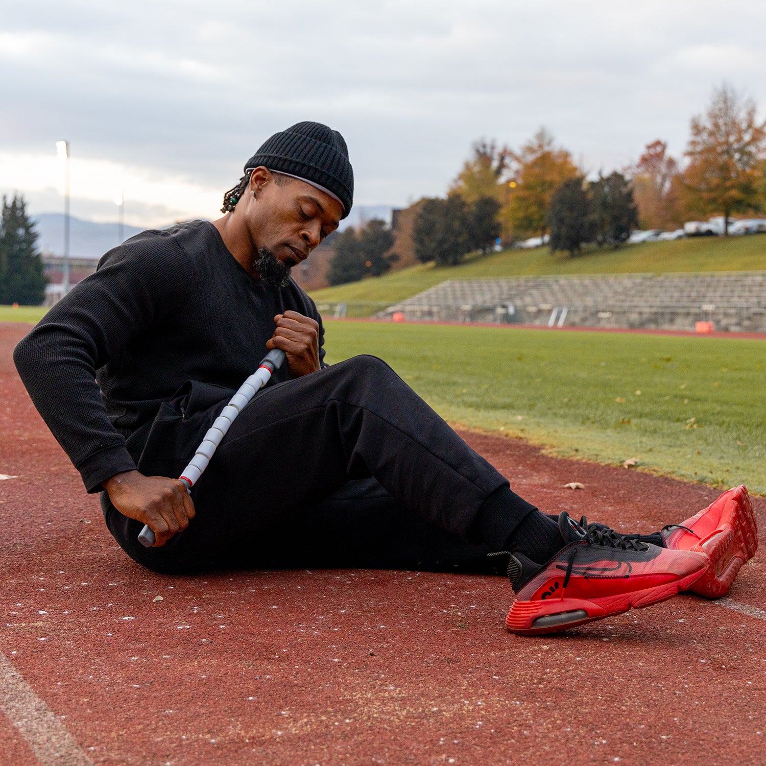 A man in black athletic wear and red sneakers sits on a running track, using the Sprinter Stick-22 on his thigh for deep tissue activation. Bleachers, grass, and autumn trees appear in the background under a cloudy sky.