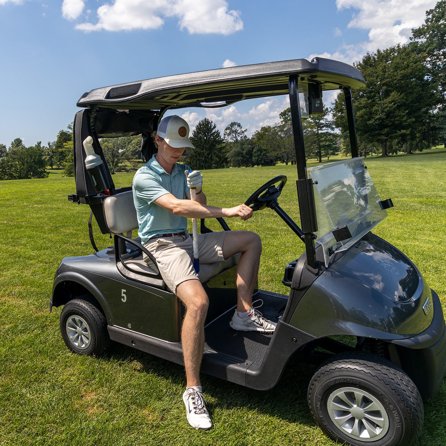 A man in a light blue shirt, shorts, and cap sits in a black golf cart on a sunny golf course, using the Body Stick- 24 for back muscle massage with trees and blue sky in the background.