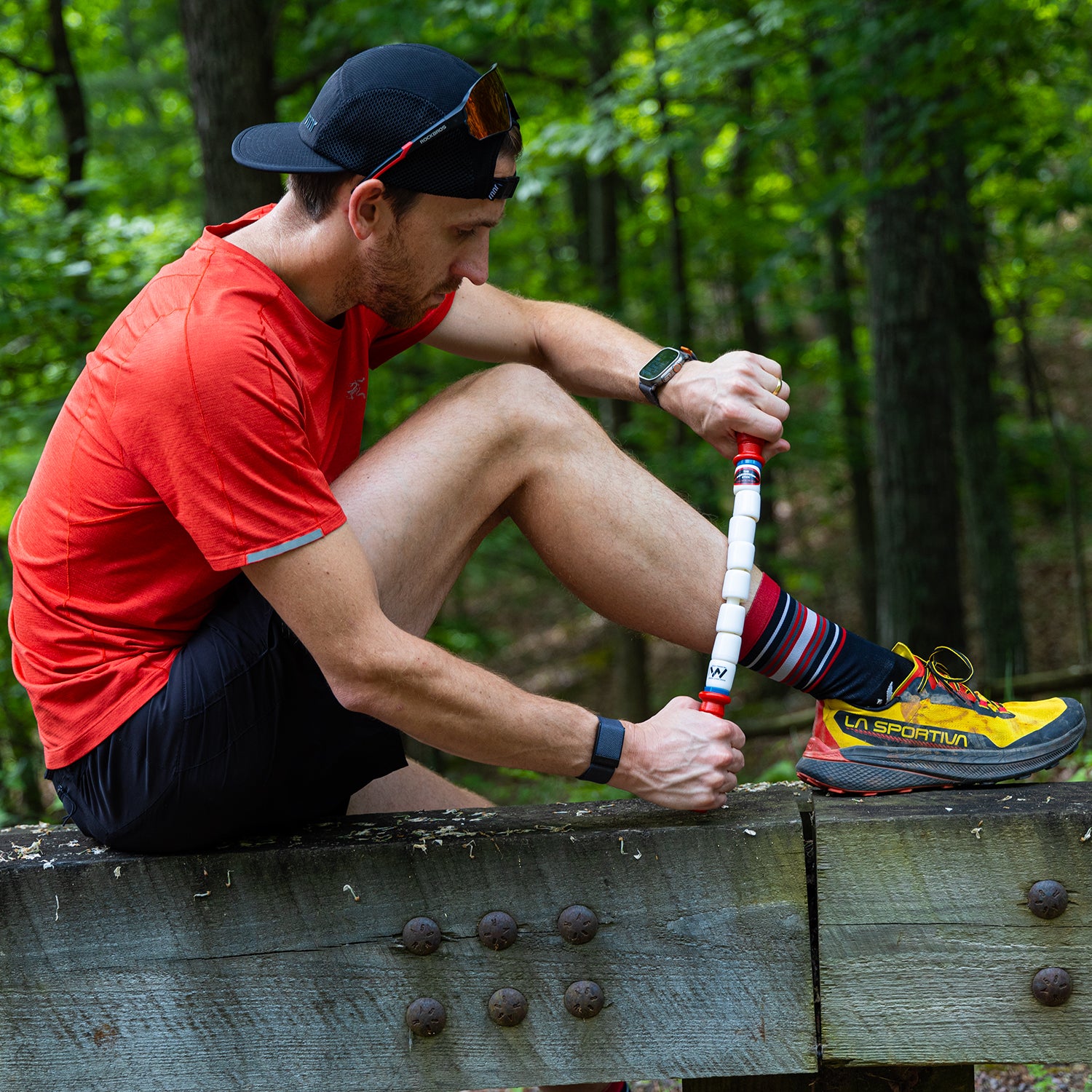 A man in athletic gear sits on a wooden beam in the forest, using his 18 Travel Stick for muscle recovery on his lower leg. He wears a red shirt, black shorts, striped socks, yellow trail shoes, and a black cap—an essential for any gym bag.