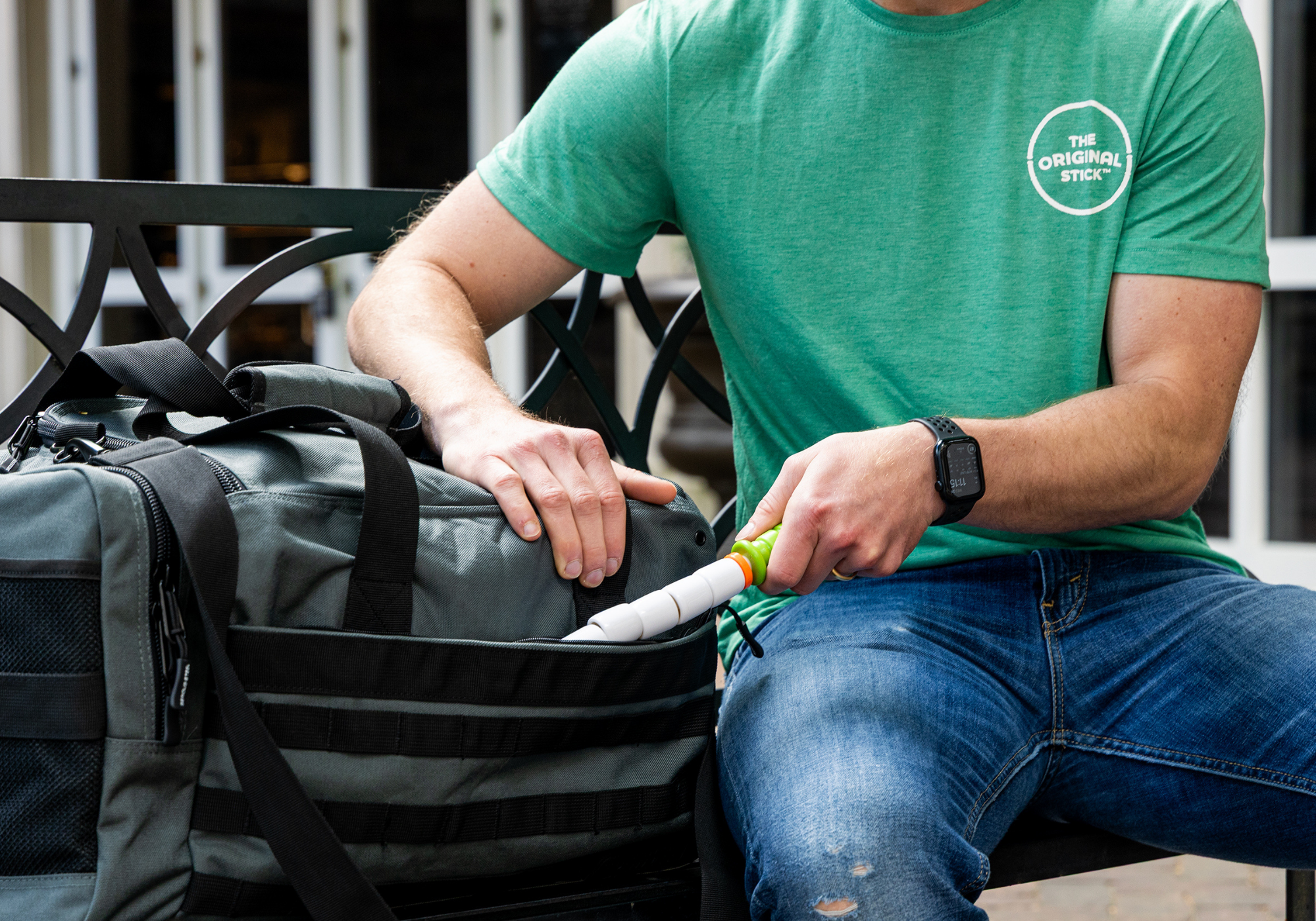 Person wearing a green Original Stick shirt taking out their Little Stick from gym bag while sitting on a park bench.