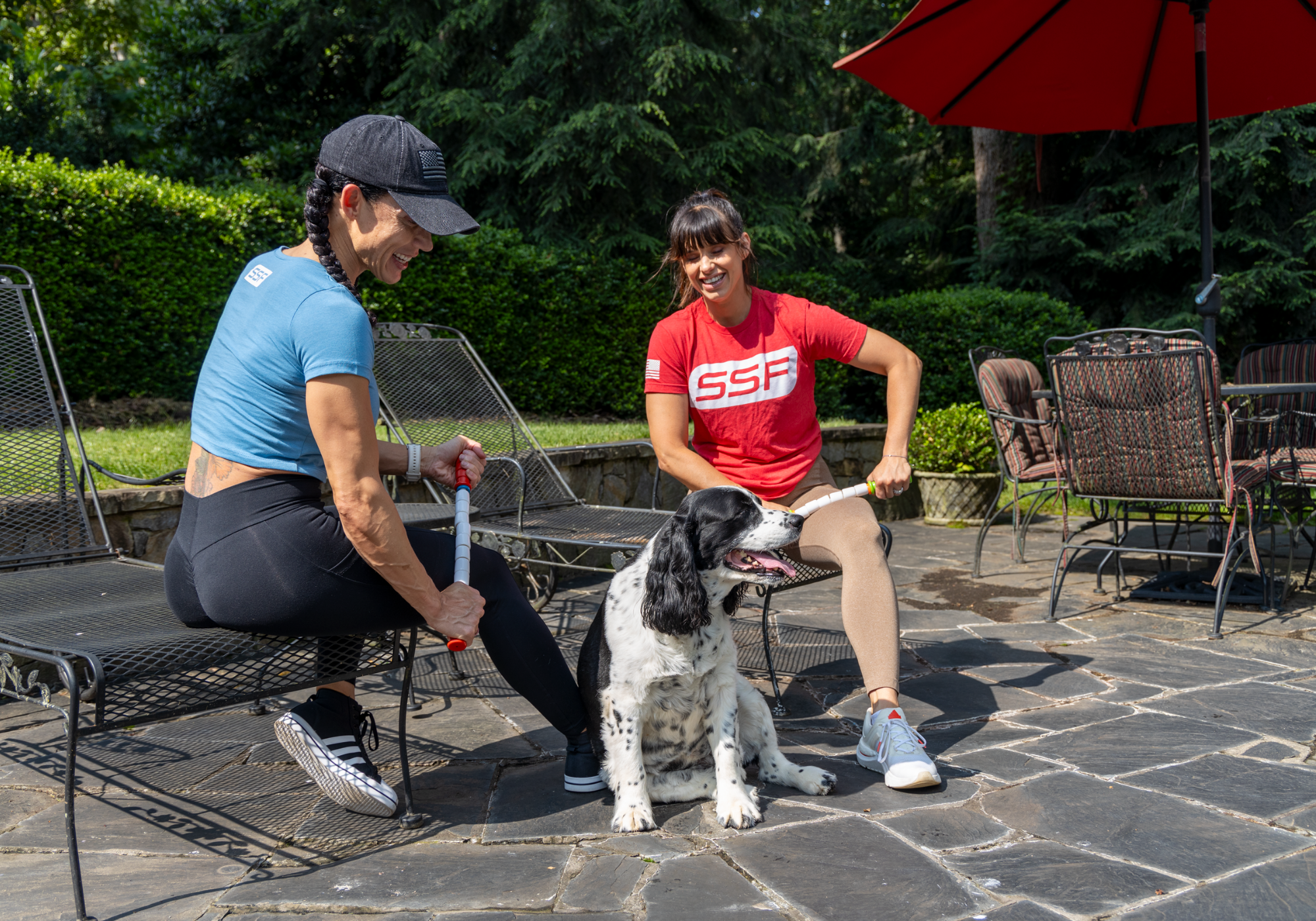 Two women sitting outside on a chairs with a dog in the sun. One is wearing a blue shirt using the Travel Stick to massage her outer quads, the other is wearing a red shirt while using the Little Stick to massage her inner quad.