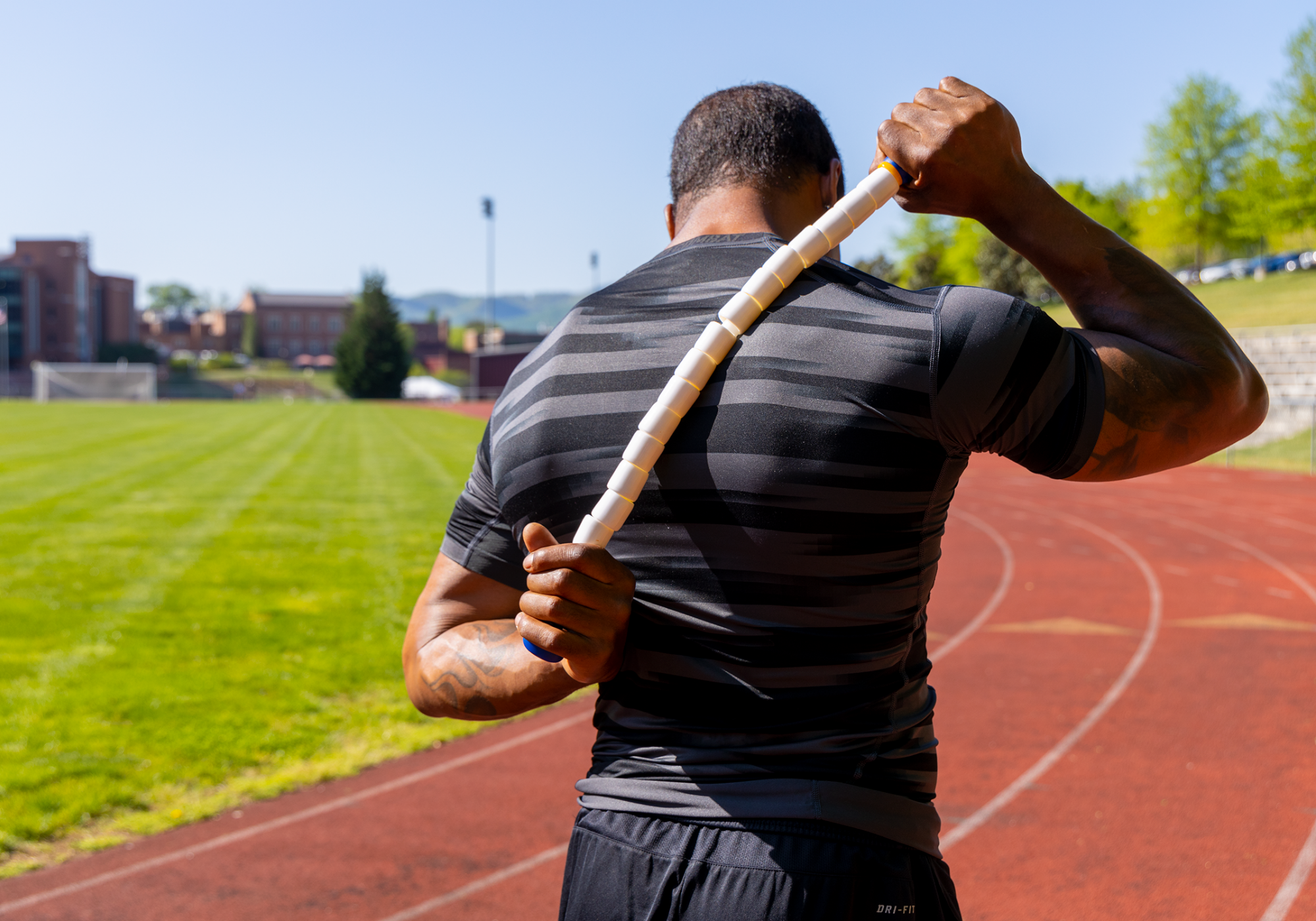 Athletic male outside standing on a track using the Body Stick to massage their upper back to warm up before a track workout.