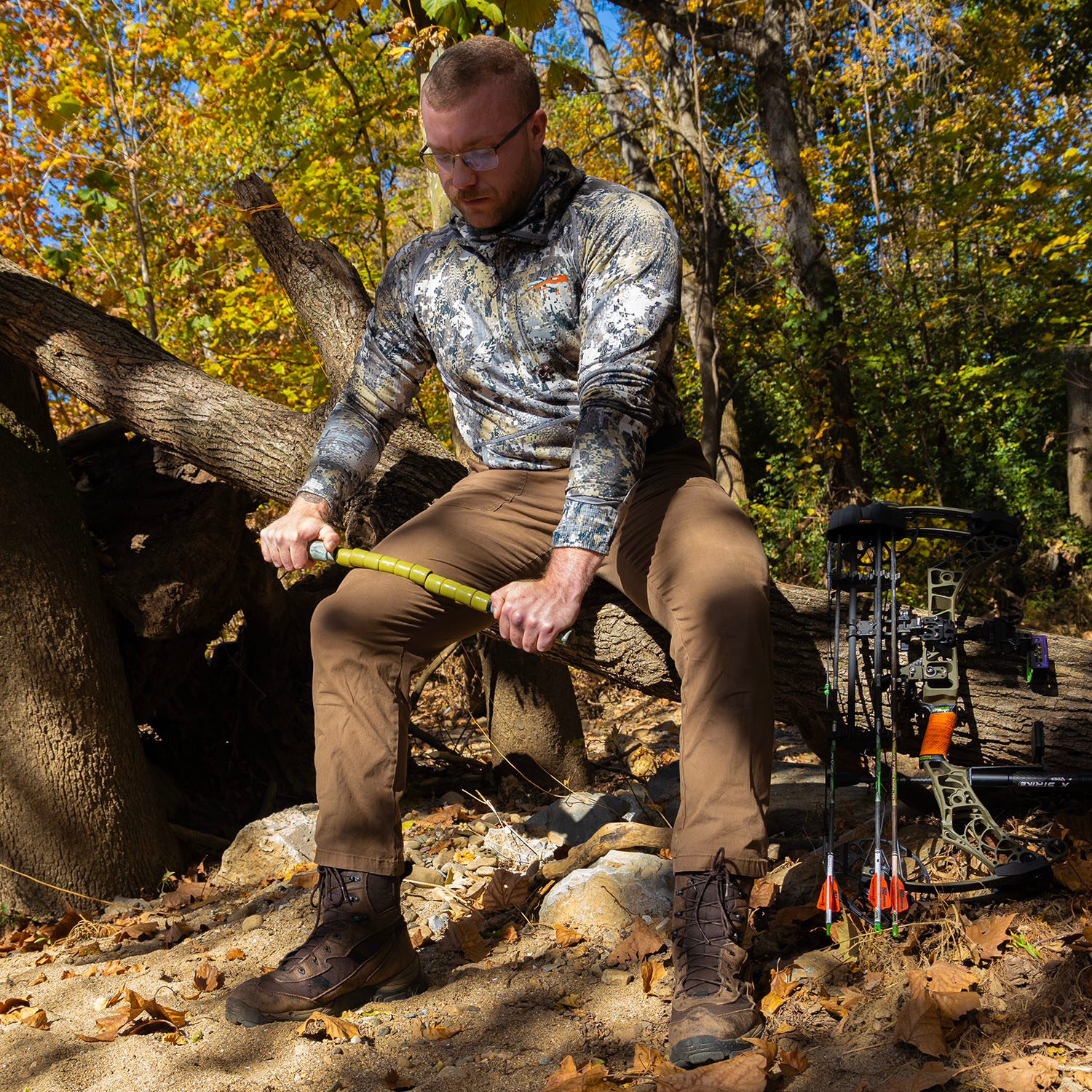Man wearing a camoflauge longsleeve and brown pants while seated on a log using the Hunter Stick to massage his right quad with a compound bow next to him.
