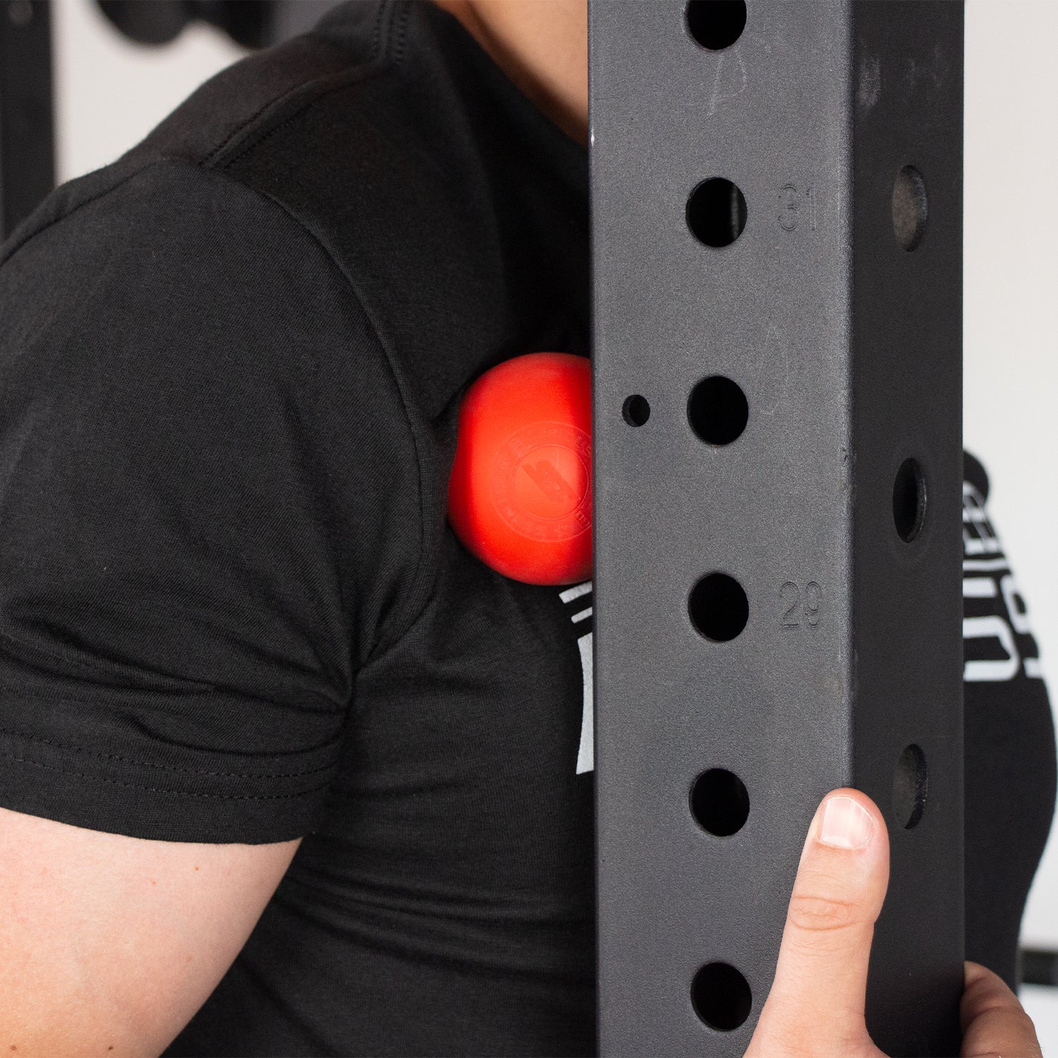 A person uses a red Lacrosse Ball against their upper chest, pressing it between their body and a black metal exercise rack with numbered holes to support mobility and recovery.