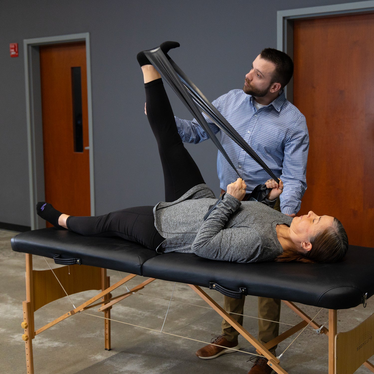 A physical therapist helps a woman lying on a massage table by stretching her raised leg with Physical Therapy Exercise Bands - 4 X 5 in a clinical setting to improve her flexibility.