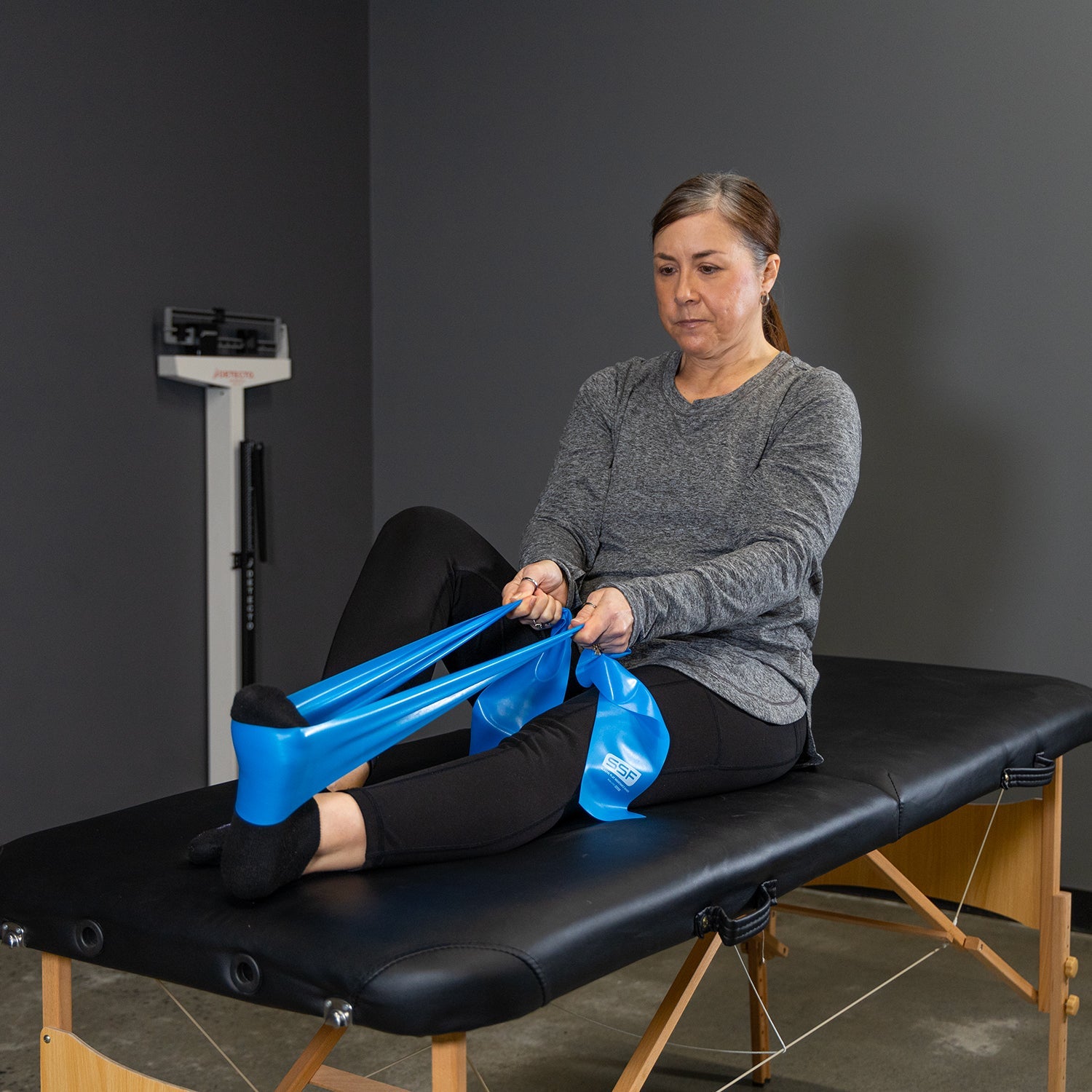 A woman sits on a black therapy table, stretching a Physical Therapy Exercise Band - 4 X 5 with her feet while holding the ends in her hands. A weight scale is visible in the background against a gray wall.