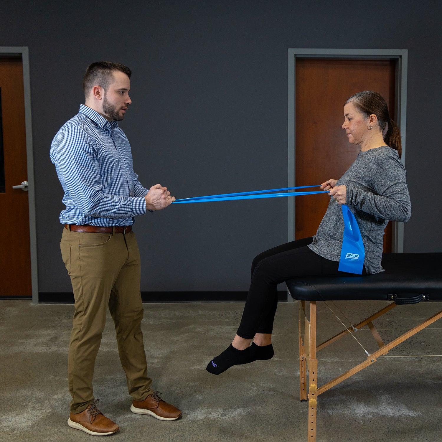 A woman sits on an exam table using Physical Therapy Exercise Bands - 4 X 5, assisted by a man, to perform flexibility exercises in a clinical setting.
