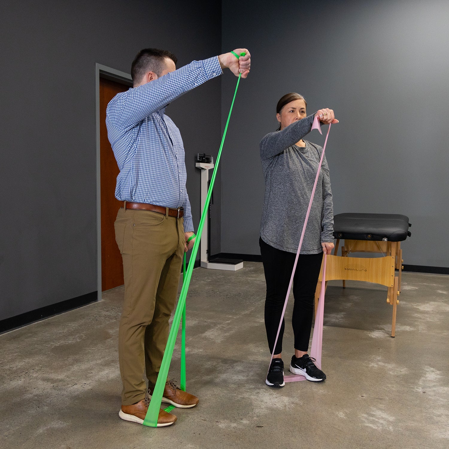 Two people use Physical Therapy Exercise Bands - 4 X 25 Yards for strength exercises, lifting their arms forward while stepping on one end of the band; a massage table appears in the background.