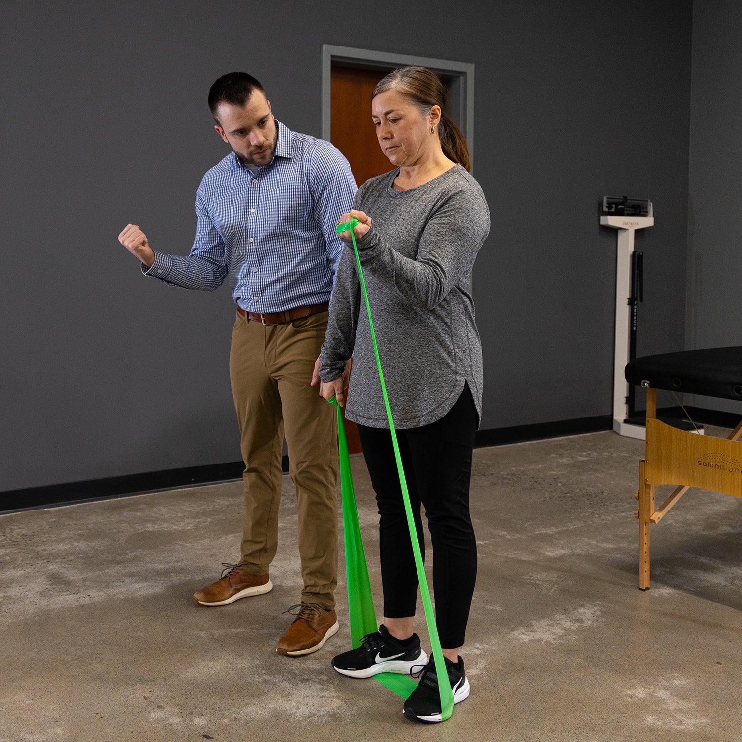 A woman uses the Physical Therapy Exercise Bands - 4 X 5 for arm exercises to improve flexibility, while a business-casual man observes and encourages her in a gym or physical therapy setting.