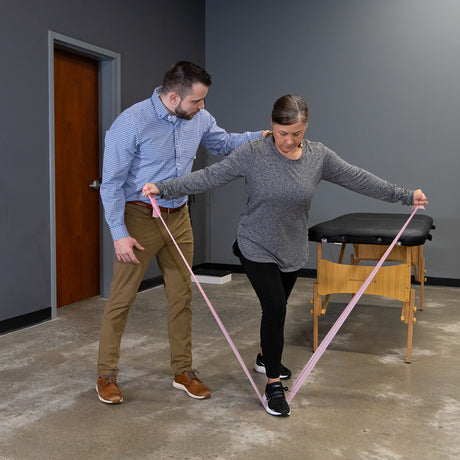A woman uses Physical Therapy Exercise Bands - 4 X 25 Yards, stepping forward with arms extended, while a man supports her in a physical therapy setting to enhance strength and flexibility.