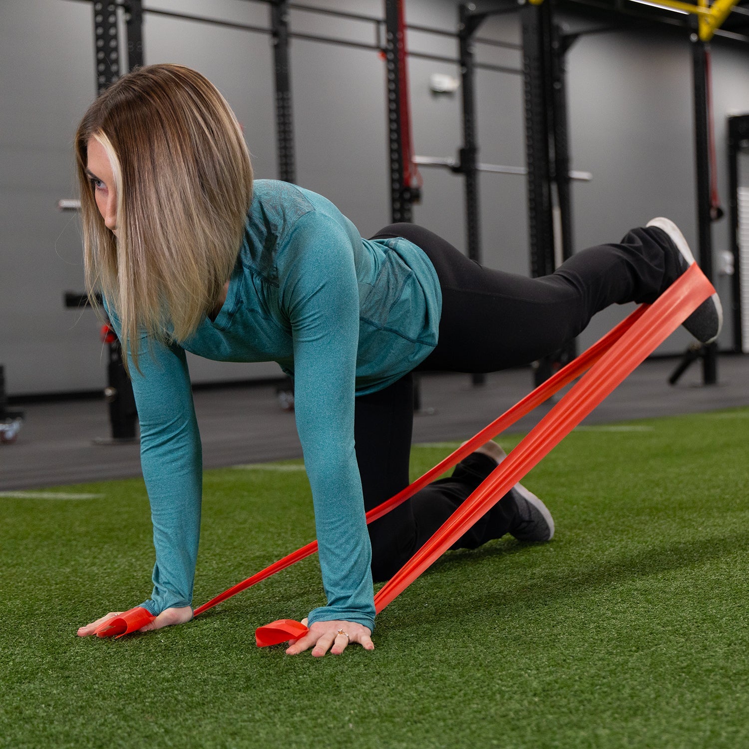 A woman in a gym uses Physical Therapy Exercise Bands - 4 X 25 Yards on artificial turf, supporting herself on hands and knees while extending one leg back for strength training.