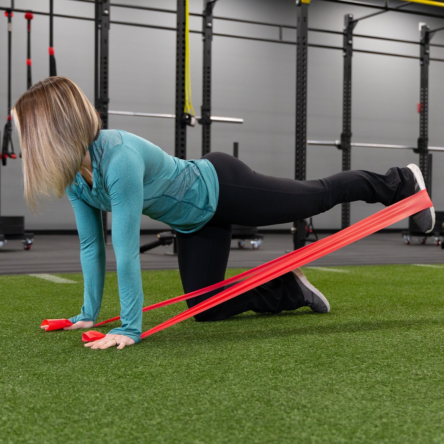 A woman in athletic wear uses Physical Therapy Exercise Bands - 4 X 5 at the gym, performing an all-fours leg extension exercise with the band anchored under her hands and foot for flexibility training.