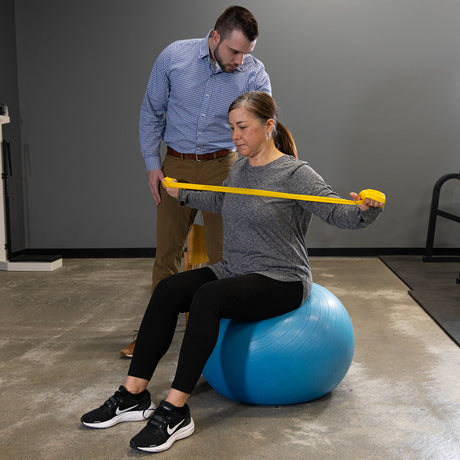 A woman stretches a Physical Therapy Exercise Band (4 X 5) while sitting on a blue exercise ball, supervised by a man in a gym or home workout setting to improve flexibility.