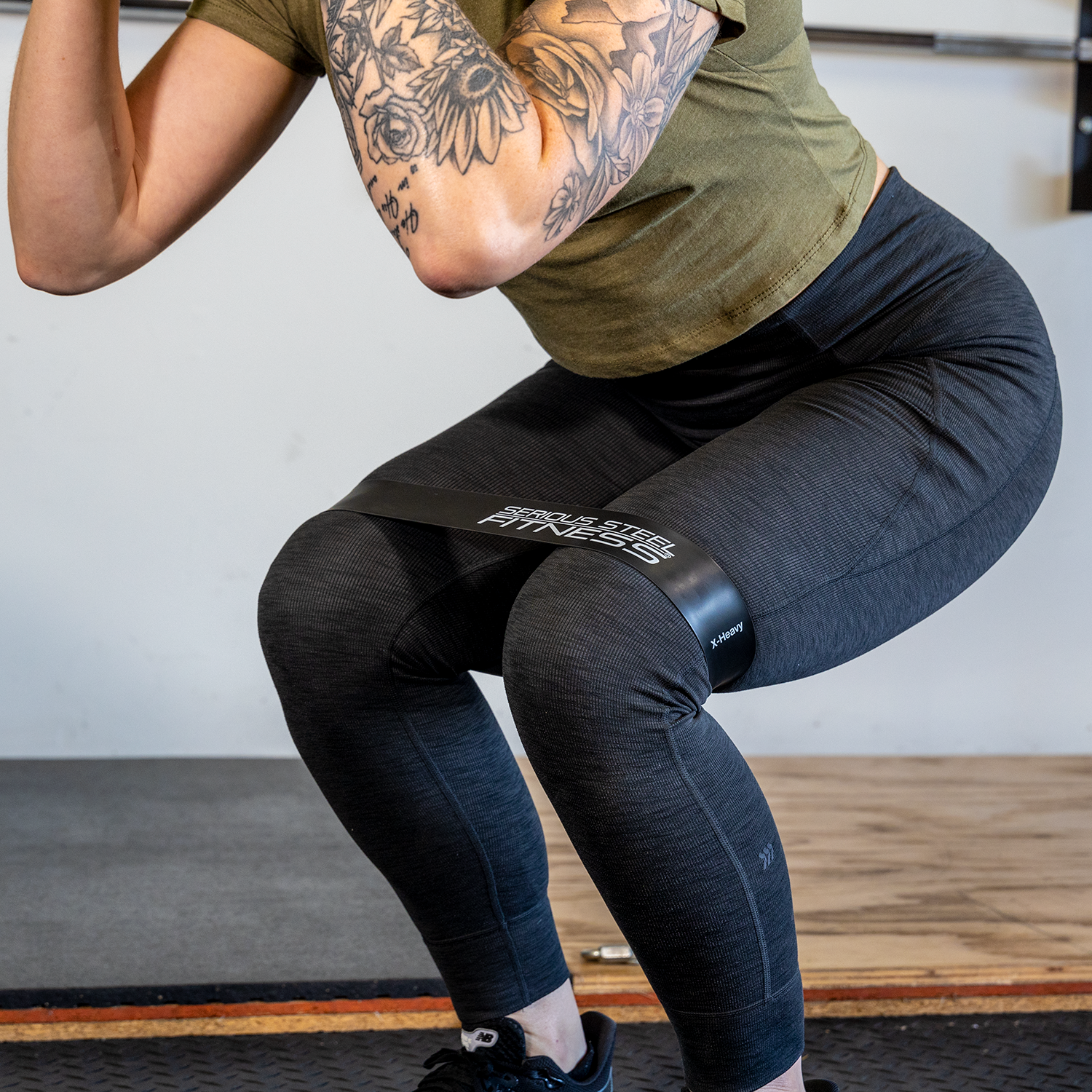 A person in a green shirt and black leggings performs squats with Mini Loop Fitness Bands - 10 Pack around their thighs, arms bent to reveal tattoos, demonstrating focused fitness band exercises in a gym setting.