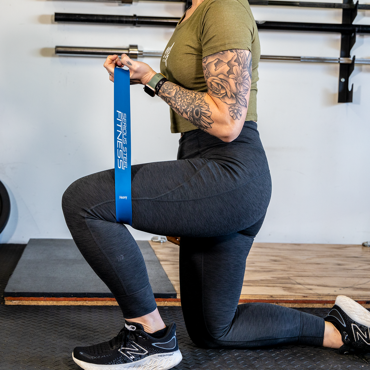 A woman in athletic wear kneels in a gym, holding a blue Mini Loop Fitness Band from the 10 Pack. Barbells are mounted on the wall behind her, and she has a floral tattoo on her arm.