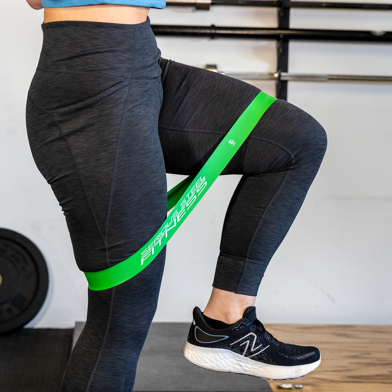 Wearing black athletic leggings and sneakers, a person performs a leg exercise in the gym using green Mini Loop Fitness Bands - 10 Pack around their thighs.