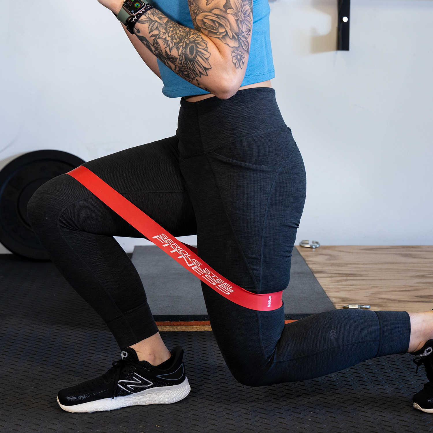 A person in black leggings and sneakers performs lunges with red Mini Loop Fitness Bands - 10 Pack around their thighs. Their tattooed arms are bent, and gym flooring plus equipment can be seen in the background.