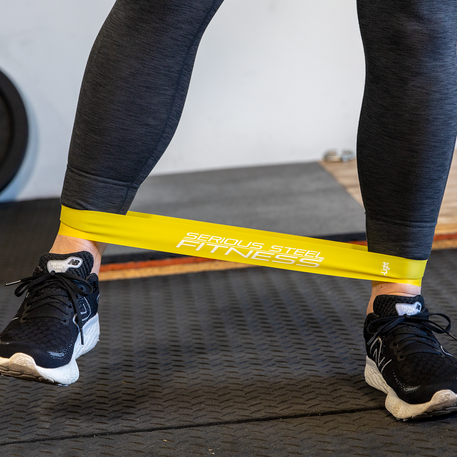 A person in black athletic shoes and dark leggings performs a resistance band exercise with a yellow Mini Loop Fitness Bands - 10 Pack band around their lower legs on the gym floor.