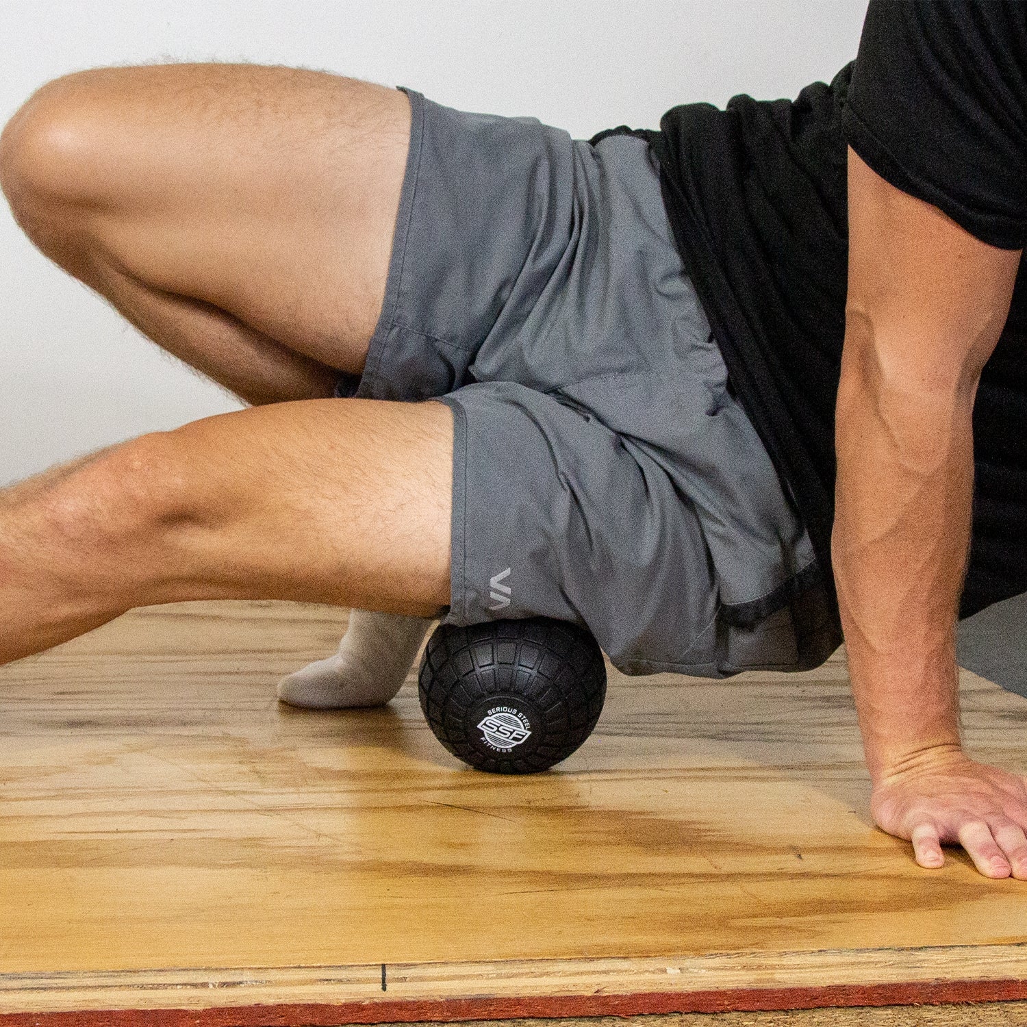 Using a 5 Massage Ball, a person in gray shorts performs deep tissue release under their thigh on a wooden floor, supporting themselves with one arm.