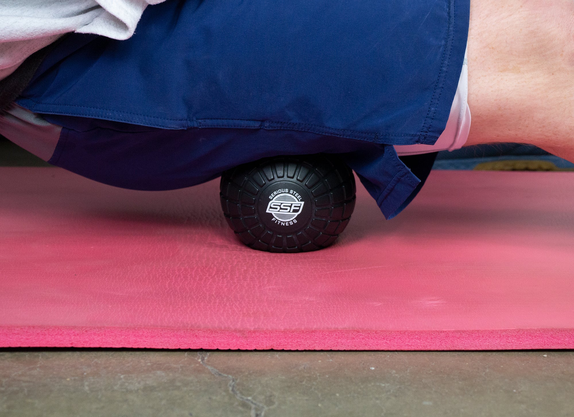 A person uses the 5 Massage Ball under their hip while lying face down on a pink mat, wearing blue athletic shorts, to demonstrate deep tissue release for better mobility.