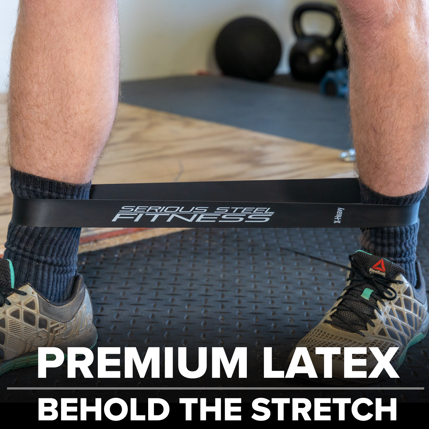 In a gym, a person stretches a black Warm-Up Resistance Band Set (2 x 12) around their legs while wearing athletic shoes. The text reads: PREMIUM LATEX BEHOLD THE STRETCH.