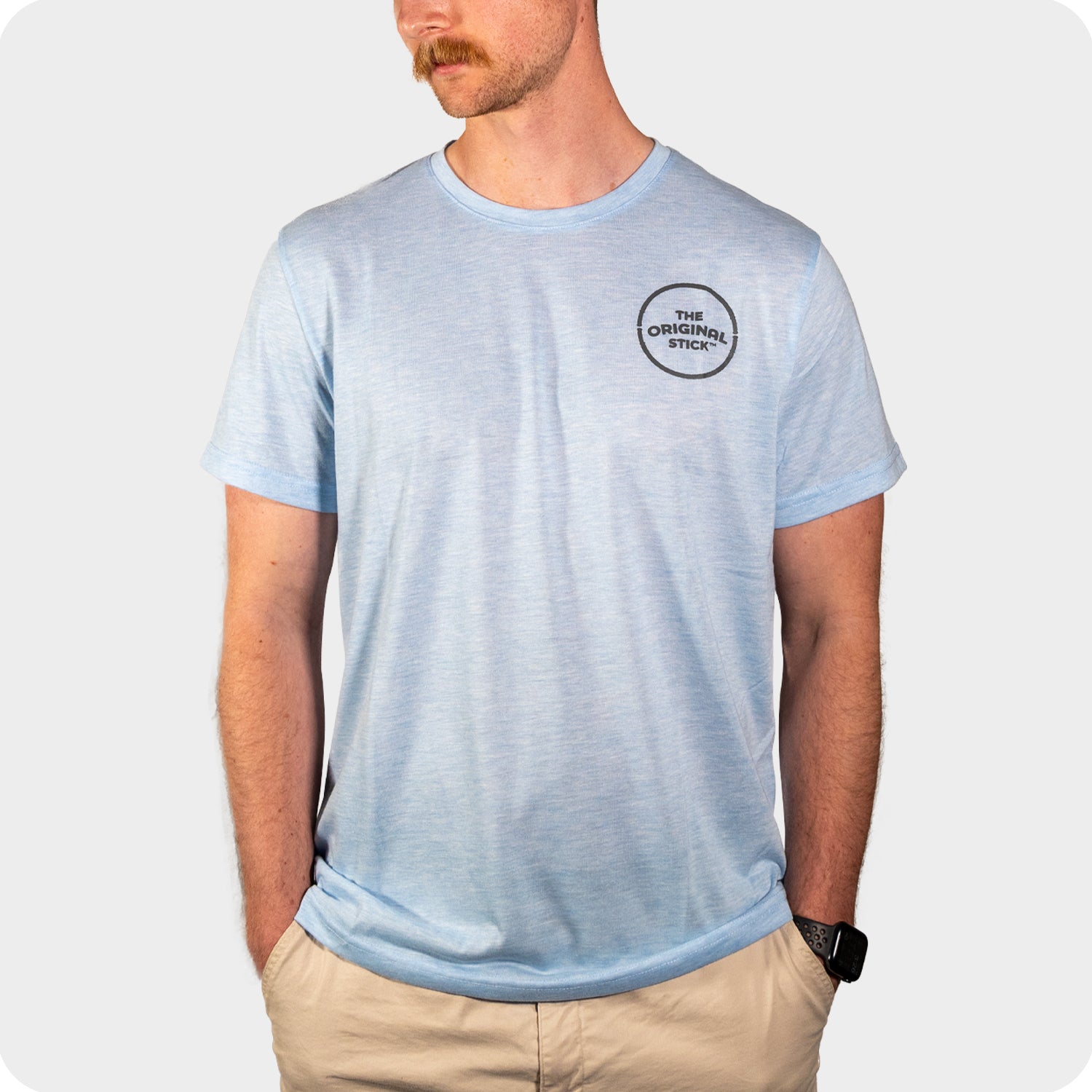 A man stands against a plain background with his hands in his pockets, wearing The Original Stick Premium Shirt—a light blue cotton-poly crew neck tee featuring THE ORIGINAL STICK logo on the left chest—and beige shorts.