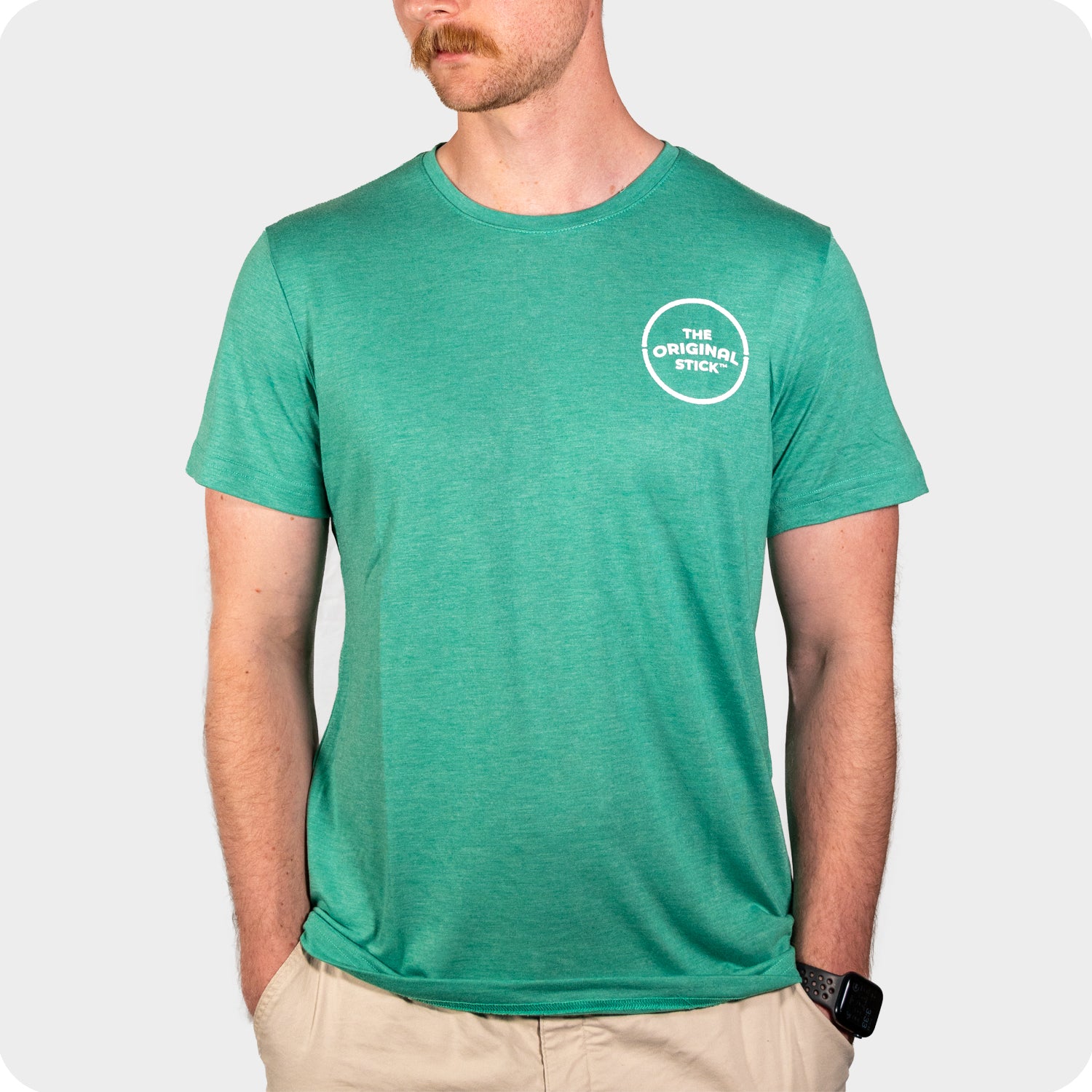 A man in The Original Stick Premium Shirt, a green crew neck with logo on the left chest, stands with hands in beige shorts pockets. He wears a mustache and smartwatch; the soft cotton-poly short sleeve shirt pops against a light gray background.
