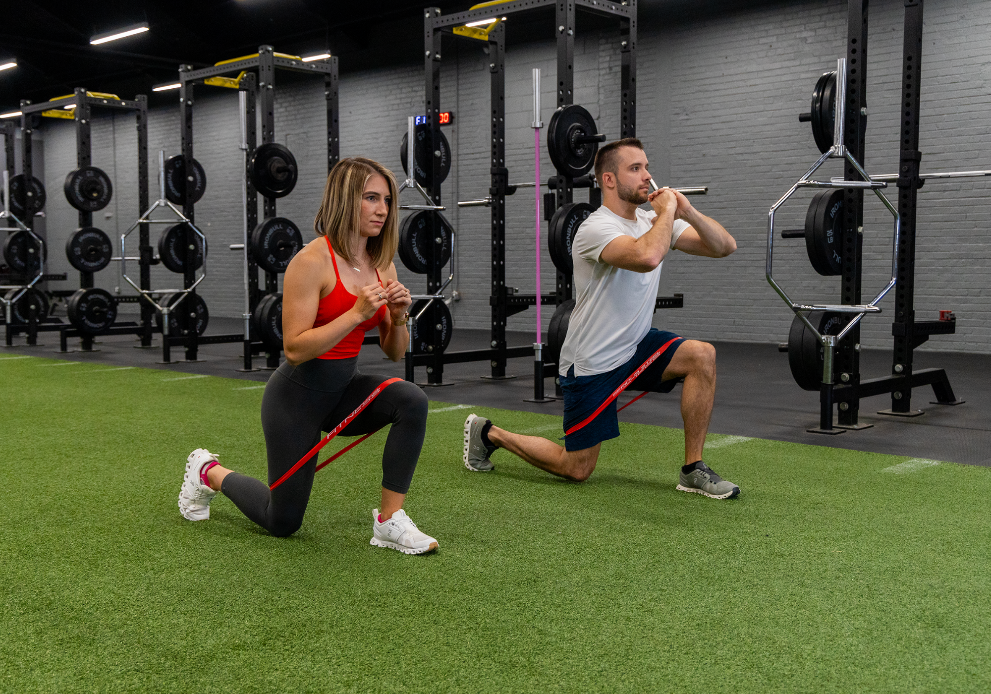Two people kneels on a gym floor, one wearing black leggings and a red top, the other wearing a white shirt and black shorts using a red resistance band around their thighs for exercise. 