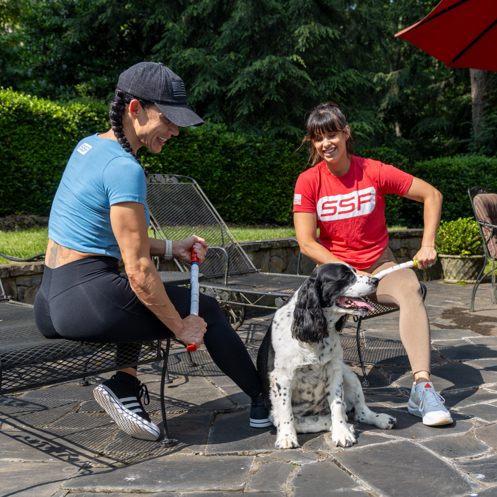 Two women sitting outside on a chairs with a dog in the sun. One is wearing a blue shirt using the Travel Stick to massage her outer quads, the other is wearing a red shirt while using the Little Stick to massage her inner quad.