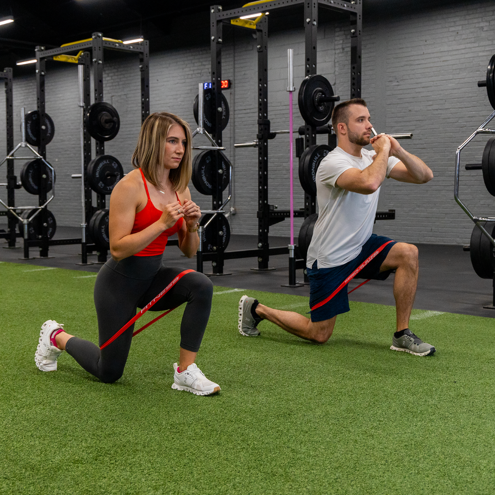Two people kneels on a gym floor, one wearing black leggings and a red top, the other wearing a white shirt and black shorts using a red resistance band around their thighs for exercise. 